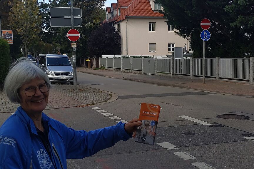 Eine Frau steht an einer Einbahnstraße und hält einen Flyer hoch, der auf die Gefahren des Linksradelns aufmerksam macht.